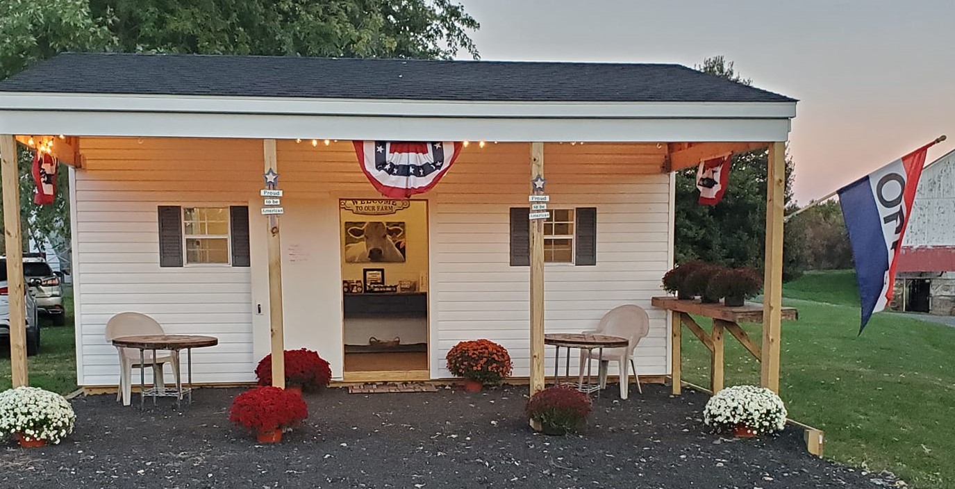 Charming farm stand with a welcoming entrance, featuring patriotic decorations, potted mums, and outdoor seating, set against a picturesque rural backdrop.