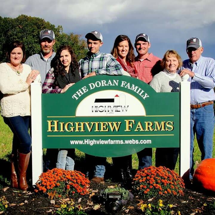 Family portrait of the Doran family in front of the Highview Farms sign, showcasing a close-knit agricultural family. The group stands outdoors with vibrant orange flowers and pumpkins in the foreground, highlighting a sunny day on the farm.